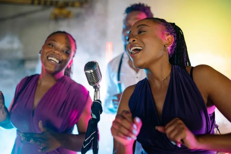 Deux jeunes femmes chantent en riant devant un micro rétro, sous des lumières roses et violettes