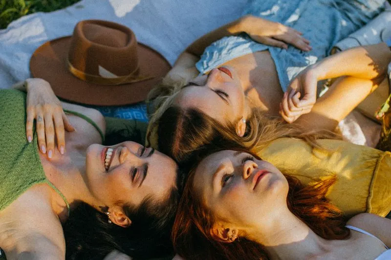 Trois jeunes femmes allongées tête contre tête sur une couverture dans l'herbe, au soleil de fin de journée