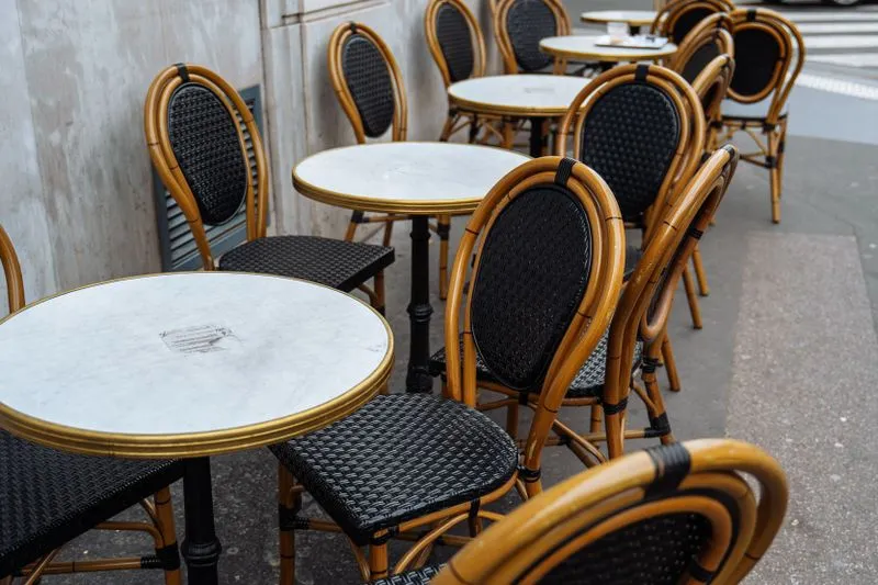 Tables rondes en marbre cerclées de laiton et chaises bistrot en rotin tressé alignées sur le trottoir d'une terrasse parisienne