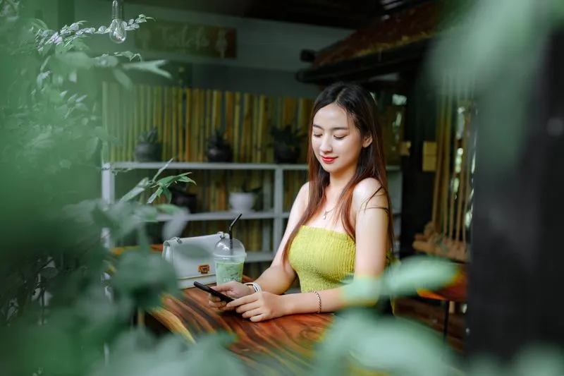 Jeune femme attablée dans un café entouré de plantes, qui regarde son smartphone, une boisson glacée posée à côté