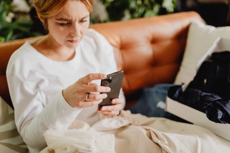 Femme en pull blanc assise sur un canapé en cuir, les yeux rivés sur l'écran de son smartphone