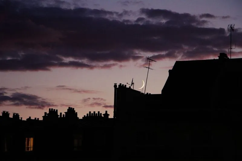 Fin croissant de lune entre les cheminées et les antennes des toits d'une ville, au crépuscule
