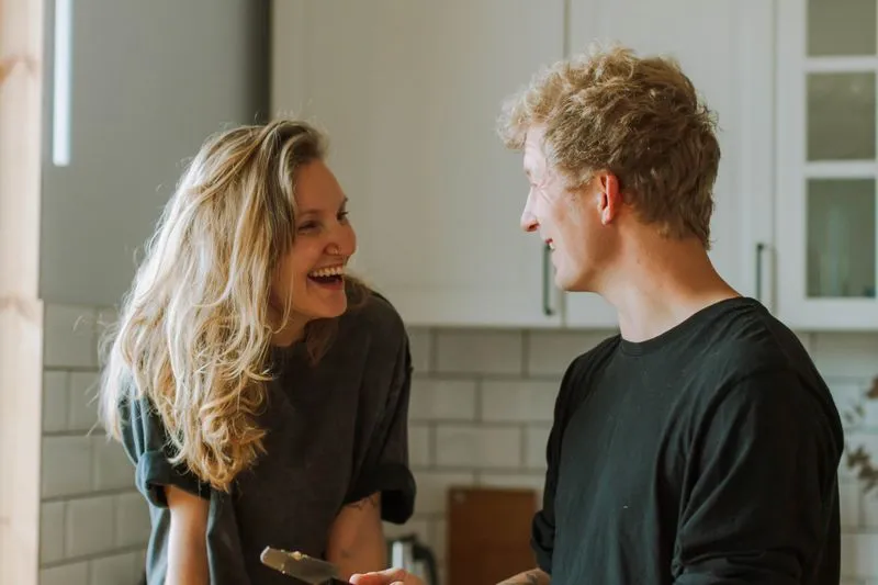 Une femme et un homme qui rient face à face dans leur cuisine, un couteau à la main