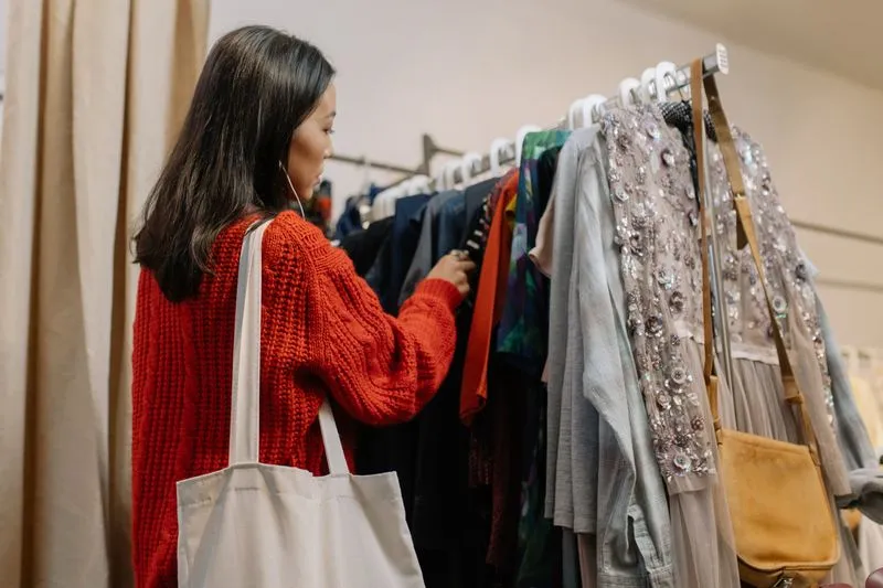 Jeune femme en pull rouge, un sac en toile à l'épaule, qui passe en revue les vêtements d'un portant de friperie
