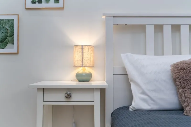 Lampe de chevet allumée sur une table de nuit blanche, à côté d'un lit et de son oreiller