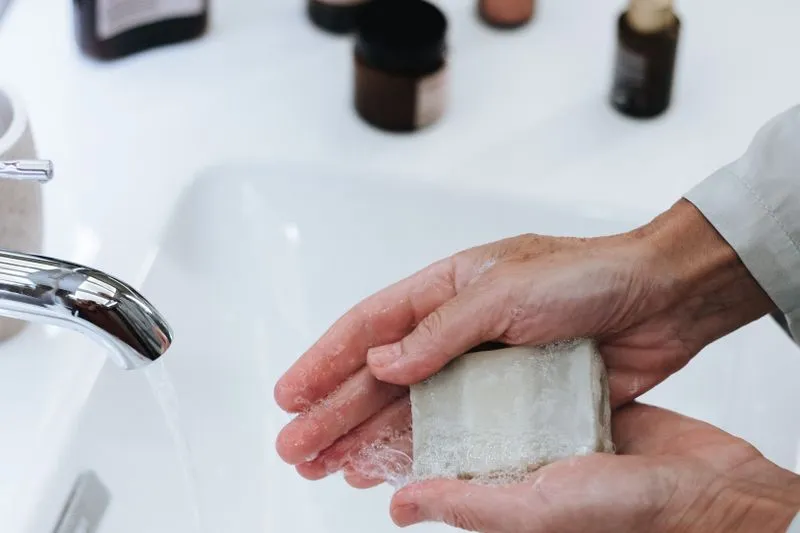 Mains qui font mousser un pain de savon sous le filet d'eau d'un robinet, au-dessus d'un lavabo blanc