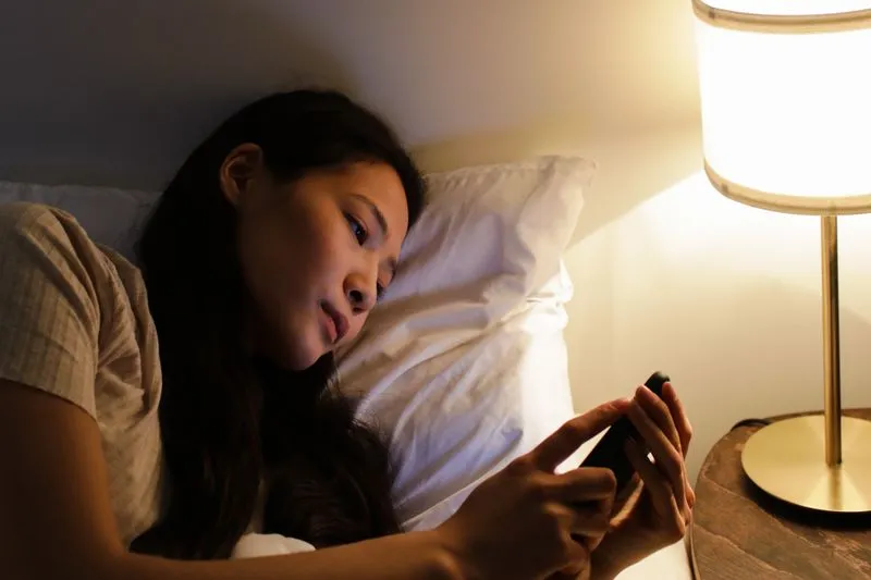 Jeune femme allongée dans son lit le soir, les yeux sur son téléphone, à côté d'une lampe de chevet allumée