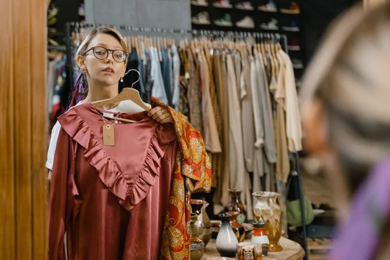 Jeune femme à lunettes qui tient contre elle une blouse unie vieux rose, face au miroir d'une friperie