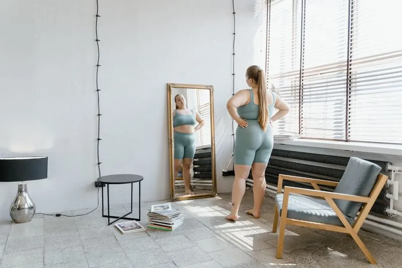 Femme debout face à un grand miroir doré posé contre le mur, à côté d'une fenêtre ensoleillée