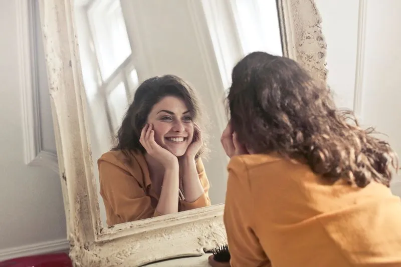 Jeune femme en haut moutarde qui se sourit dans un grand miroir ancien, près d'une fenêtre
