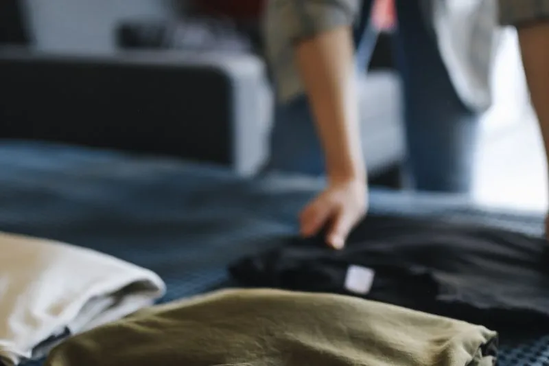 T-shirts unis pliés sur un lit, crème, kaki et noir, qu'une main est en train de ranger
