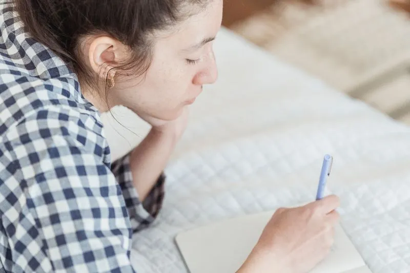 Femme en chemise à carreaux allongée sur son lit, qui écrit dans un carnet