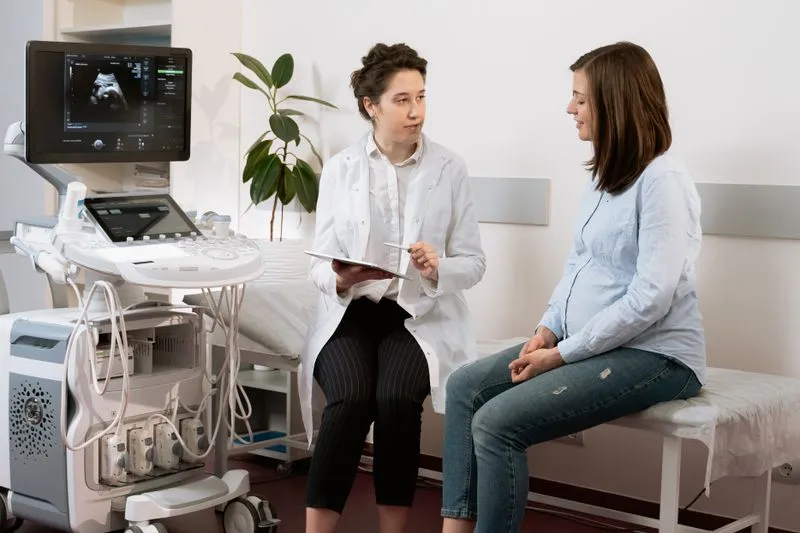 Femme enceinte assise sur la table d'examen, en discussion avec une soignante qui prend des notes, dans une salle de consultation lumineuse avec échographe