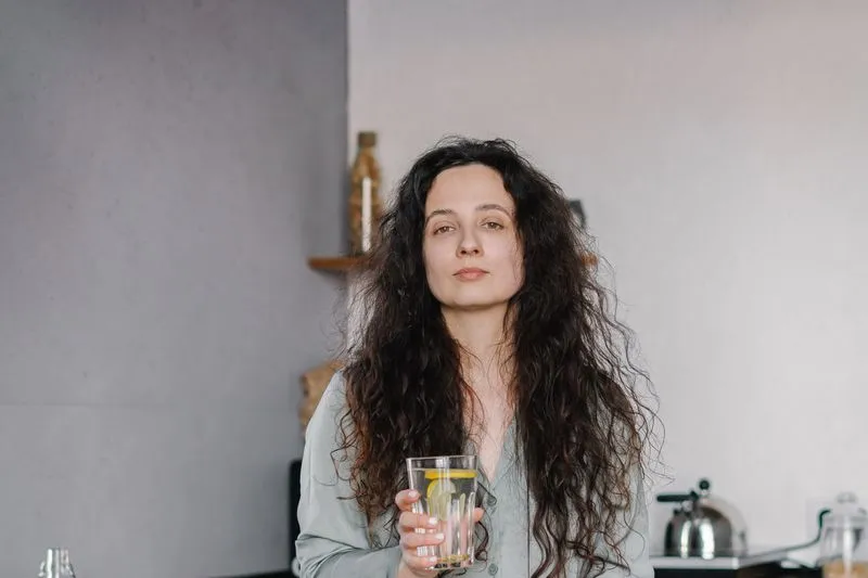 Jeune femme aux longs cheveux bouclés, en pyjama et l'air fatigué, un verre d'eau citronnée à la main, dans sa cuisine avec une bouilloire en arrière-plan