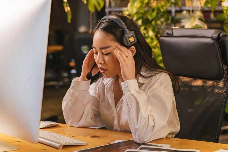 Femme qui fait une pause à son bureau, les yeux fermés, les doigts sur les tempes, devant son ordinateur