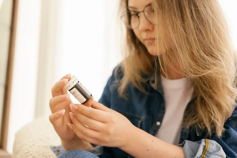 Jeune femme à lunettes qui lit l'étiquette d'un flacon de médicament, assise chez elle