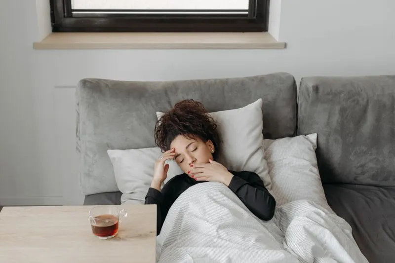 Jeune femme allongée sous une couette sur un canapé gris, les yeux fermés, une main sur le front et l'autre près de la bouche, une tasse de thé posée sur la table à côté d'elle