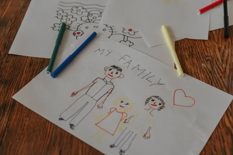 Dessin d'enfant intitulé « My Family », trois personnages au feutre sur une feuille, entourée de feutres de couleur sur une table en bois