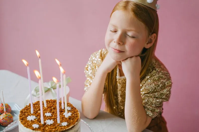 Une fillette coiffée d'une couronne dorée, les yeux fermés et le menton posé sur les mains, devant un gâteau d'anniversaire aux bougies allumées, sur un fond rose
