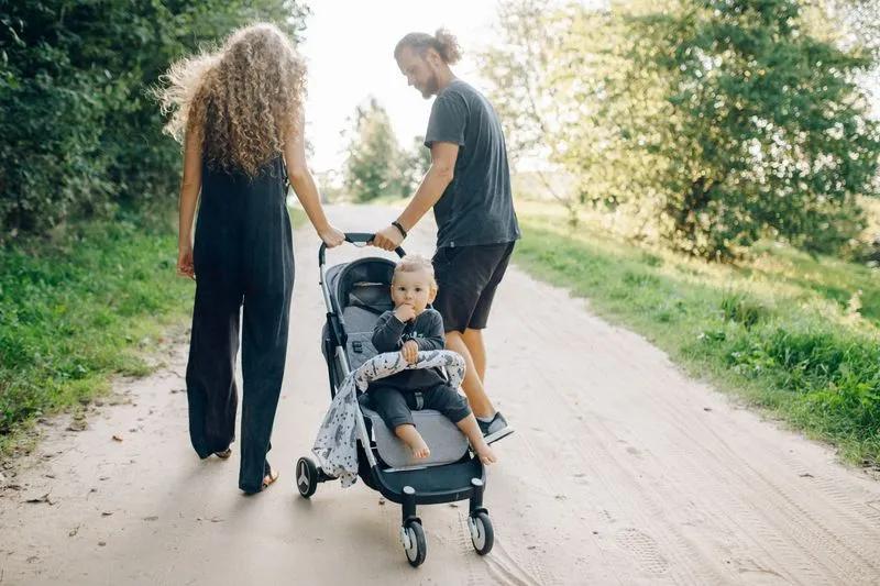 Un couple pousse une poussette sur un chemin de terre bordé d'arbres, un enfant assis à l'intérieur
