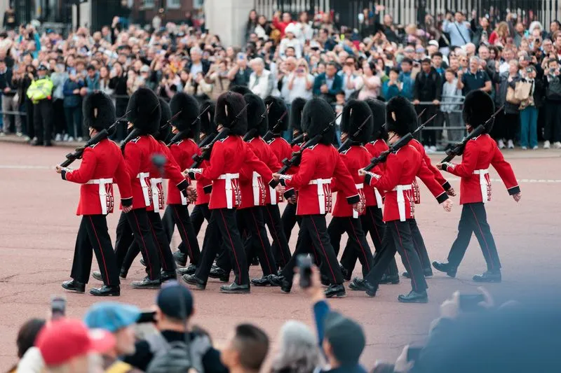 Gardes en tunique rouge et bonnet à poil défilant devant une foule, près des grilles du palais de Buckingham