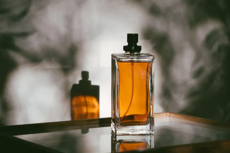 Flacon de parfum en verre rempli d'un liquide ambré, posé sur un plateau miroir dans une lumière chaude, avec son reflet