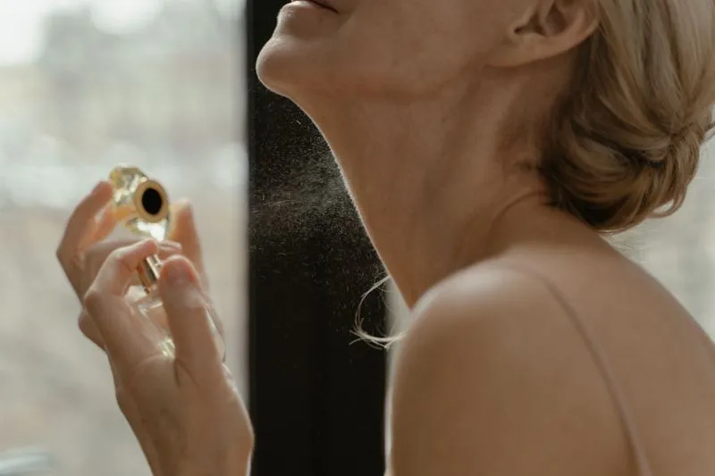 Femme vaporisant du parfum dans son cou, près d'une fenêtre lumineuse, le nuage de vaporisation visible