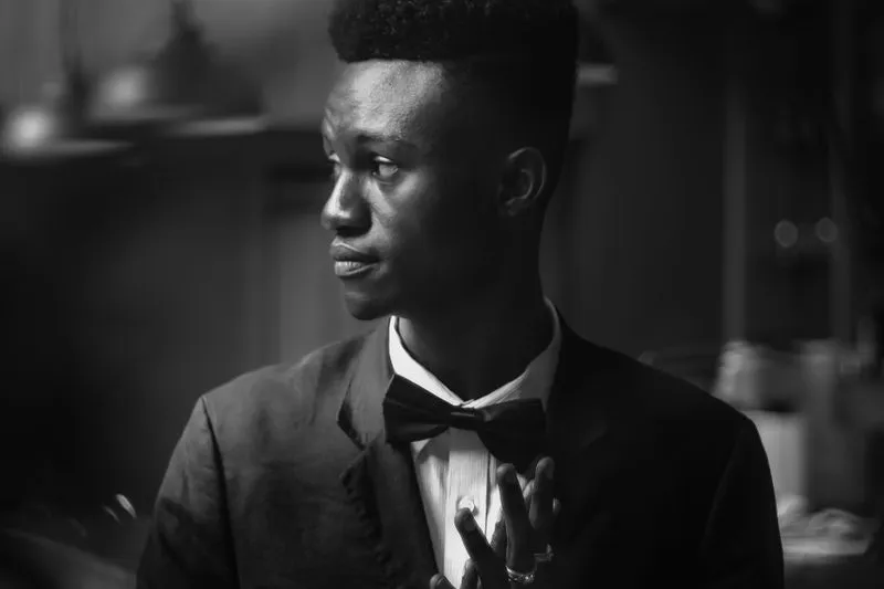 Portrait en noir et blanc d'un jeune homme noir en veste de costume sombre, chemise blanche et nœud papillon, le regard tourné de côté