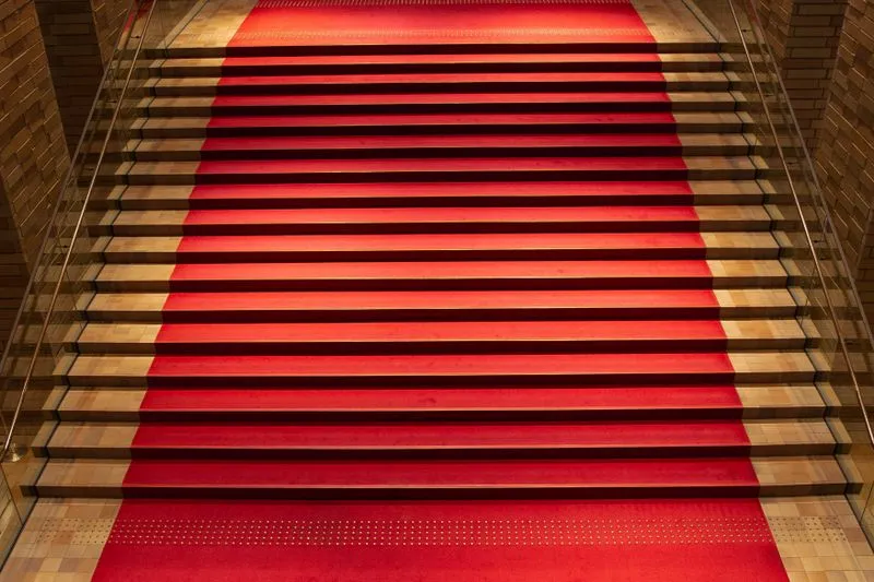 Tapis rouge déroulé sur un escalier intérieur, entre rampes de verre et lumière chaude