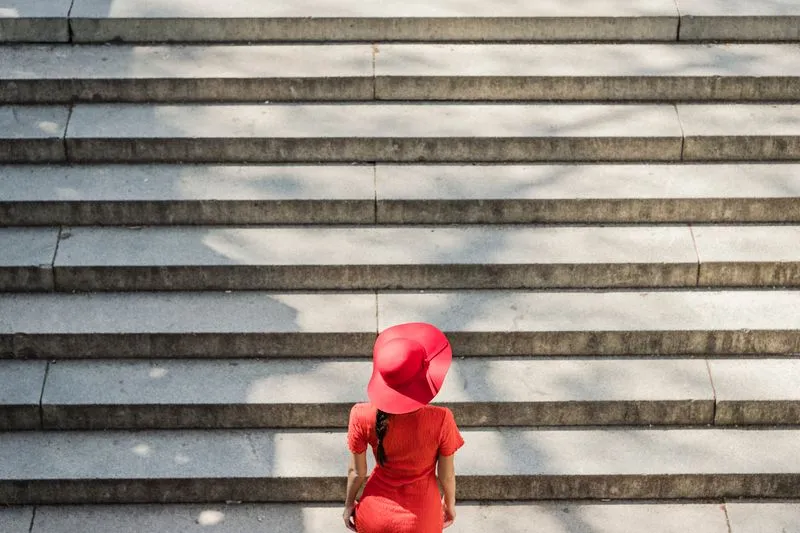 Femme en robe rouge et chapeau à large bord qui monte un grand escalier de pierre