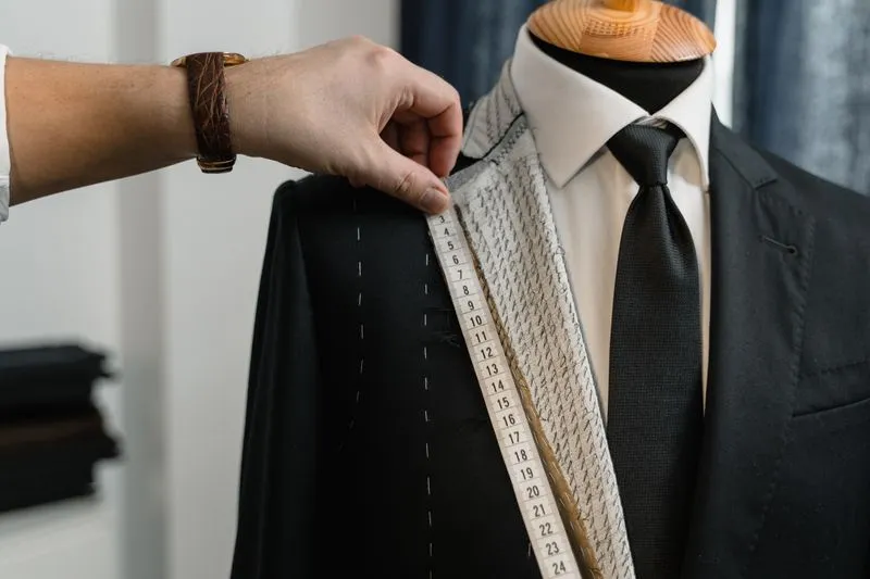 Main de tailleur qui prend les mesures d'une veste de costume noire sur un buste, chemise et cravate déjà en place