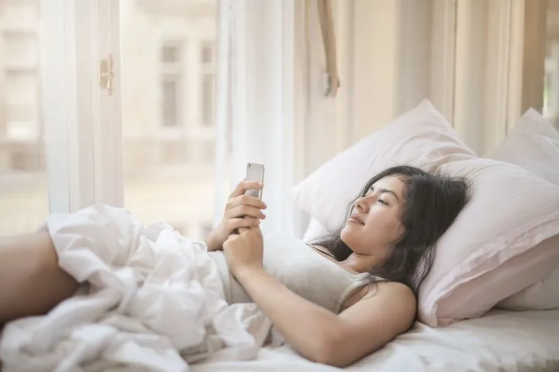 Jeune femme allongée dans son lit au petit matin, qui regarde l'écran de son téléphone