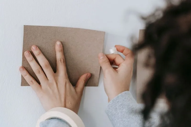 Mains qui fixent un carré de carton kraft sur un mur blanc peint avec du ruban adhésif, un rouleau de ruban de masquage au premier plan