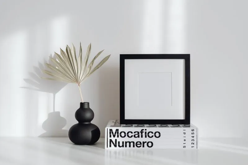 Cadre noir à passe-partout blanc posé sur un gros livre contre un mur blanc, à côté d'un vase noir garni d'une feuille de palmier séchée