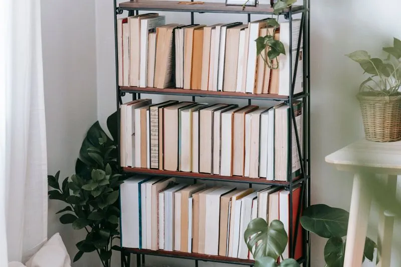 Étagère noire autoportante remplie de livres, entourée de plantes vertes en pot contre un mur blanc