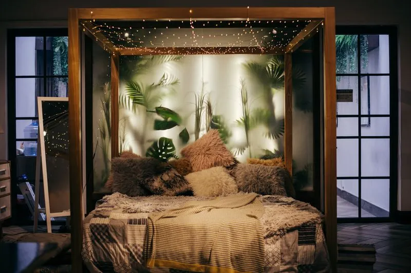Lit à baldaquin en bois la nuit, éclairé par une guirlande lumineuse tendue au-dessus et un décor de plantes en fond