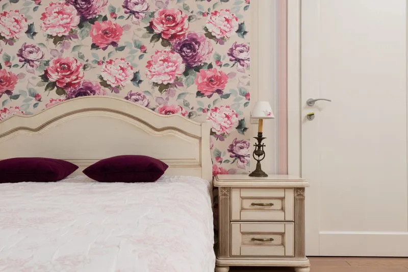 Papier peint à grosses fleurs roses et violettes sur le mur derrière une tête de lit crème, le mur voisin resté blanc autour de la porte