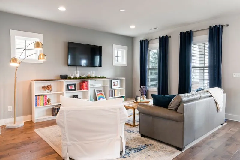 Salon lumineux avec un tapis à motifs sur un parquet, un fauteuil sous housse blanche, un canapé en cuir gris et des rideaux bleu marine