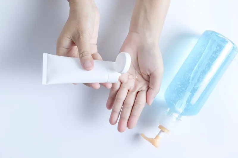 Mains pressant quelques gouttes de gel transparent d'un tube blanc sans étiquette, à côté d'un flacon pompe de gel bleuté