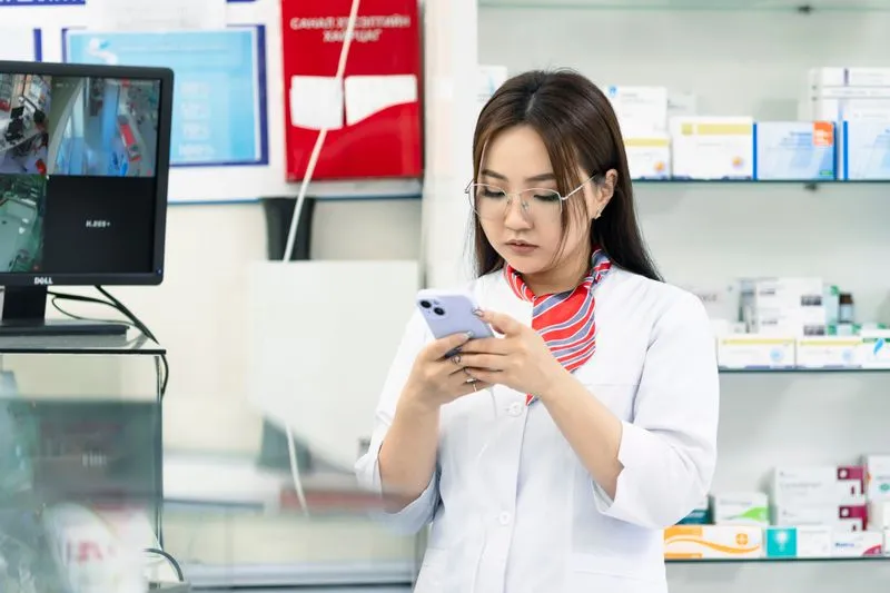 Jeune pharmacienne en blouse blanche, téléphone en main, devant des étagères de boîtes de médicaments