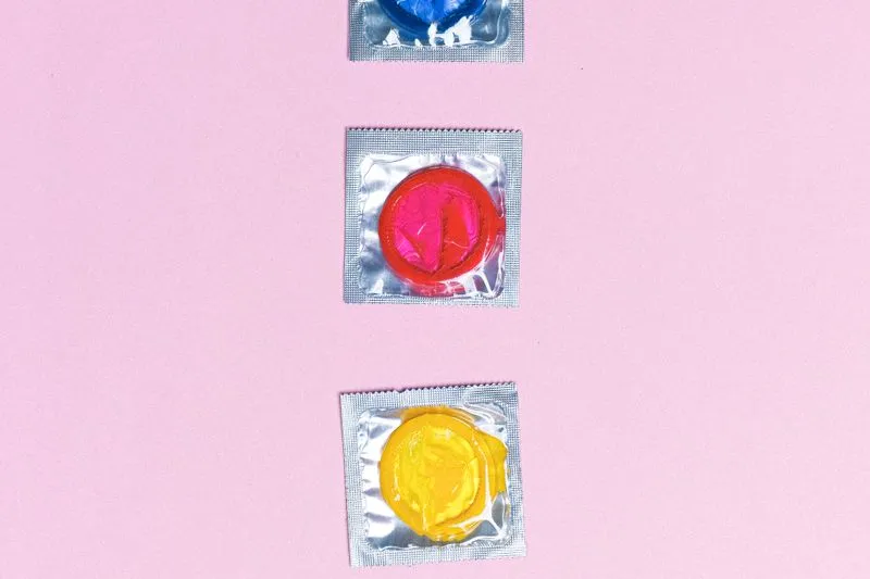 Trois préservatifs emballés, bleu, rose et jaune, alignés sur un fond rose