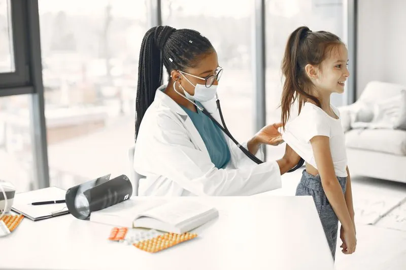 Une pédiatre ausculte avec un stéthoscope le dos d'une fillette souriante, en cabinet médical
