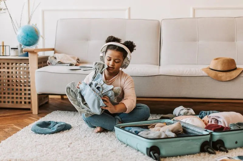 Fillette assise en tailleur, casque sur les oreilles, pliant ses vêtements devant une valise ouverte à moitié remplie