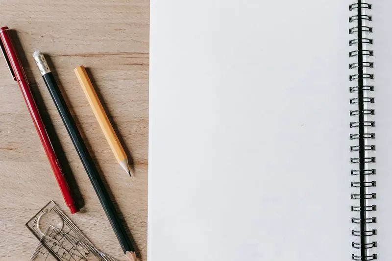 Crayons, feutre rouge et instruments de géométrie posés sur une table en bois, à côté d'un cahier à spirale ouvert sur une page blanche