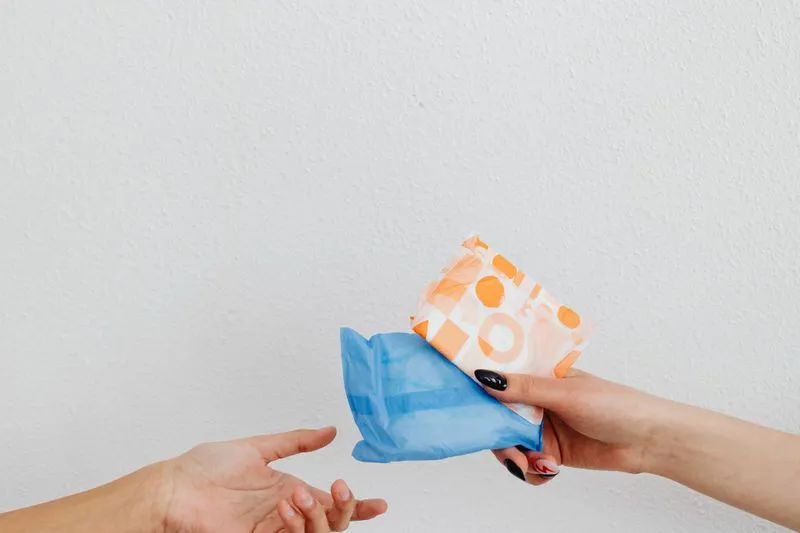 Gros plan sur deux mains : l'une tend deux serviettes hygiéniques emballées à l'autre
