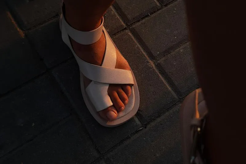 Pied d'une femme en sandale claire à brides croisées, posé sur des pavés éclairés par une lumière dorée