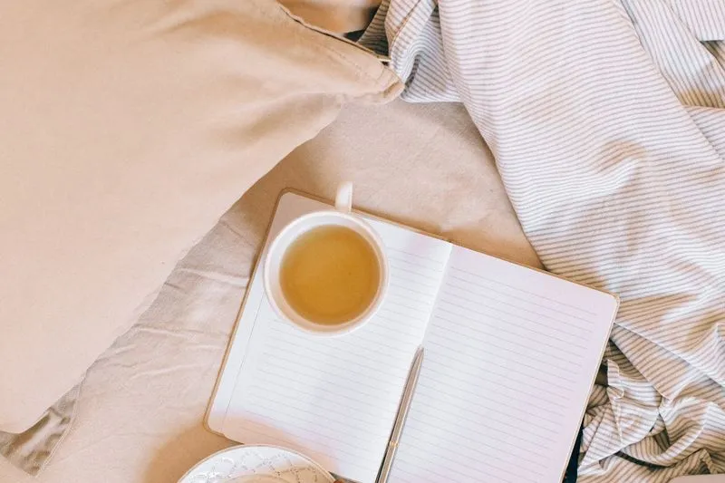 Carnet ouvert avec un stylo, posé sur un lit à côté d'une tasse de tisane