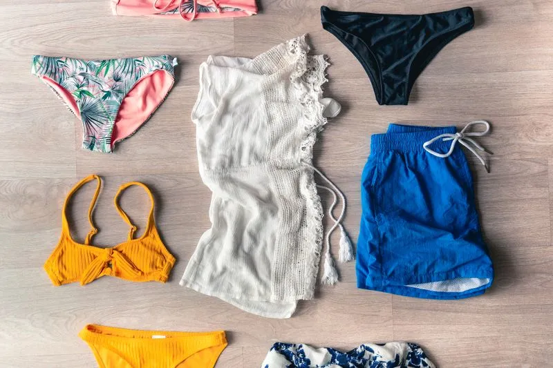 Hauts et bas de bikini de couleurs et formes différentes, un paréo en dentelle et un short de bain, disposés à plat sur un sol en bois