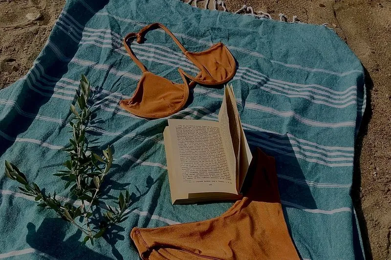 Haut et bas de bikini couleur rouille posés à plat sur une fouta rayée bleue, près d'un livre ouvert, sur le sable