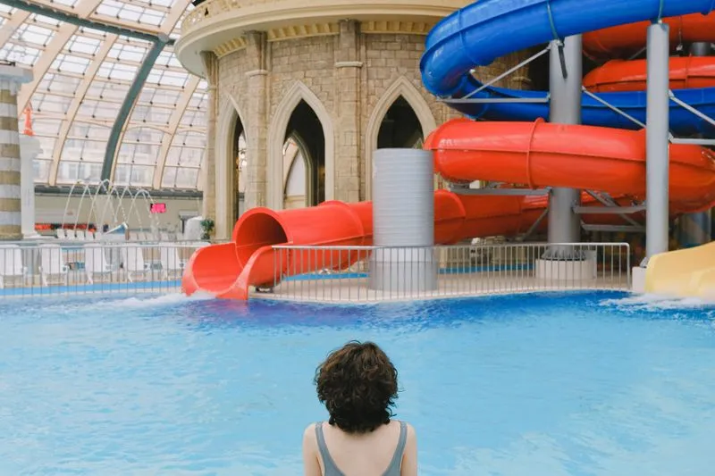 Femme de dos, bretelles de maillot gris, face aux toboggans d'une piscine intérieure